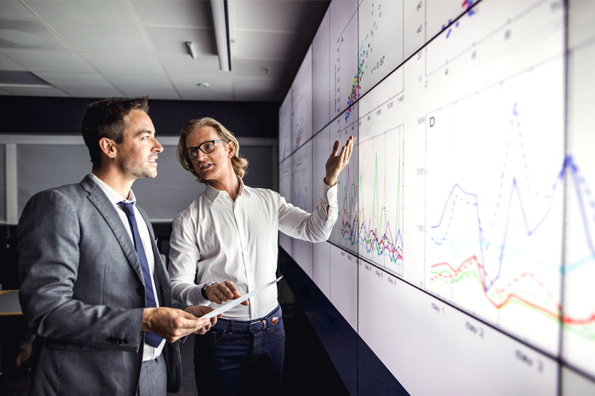 Two men in business attire look at a chart projected onto a wall. They talk as one of the men gestures to the data.